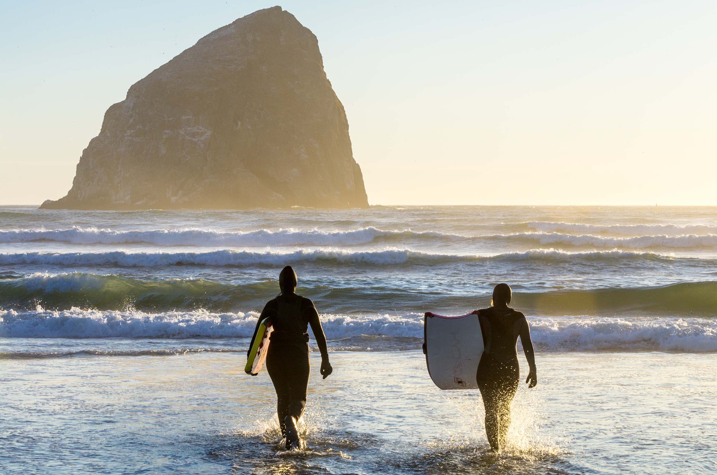 Two people walking into the ocean with surfboards in front of Haystack Rock near Headlands