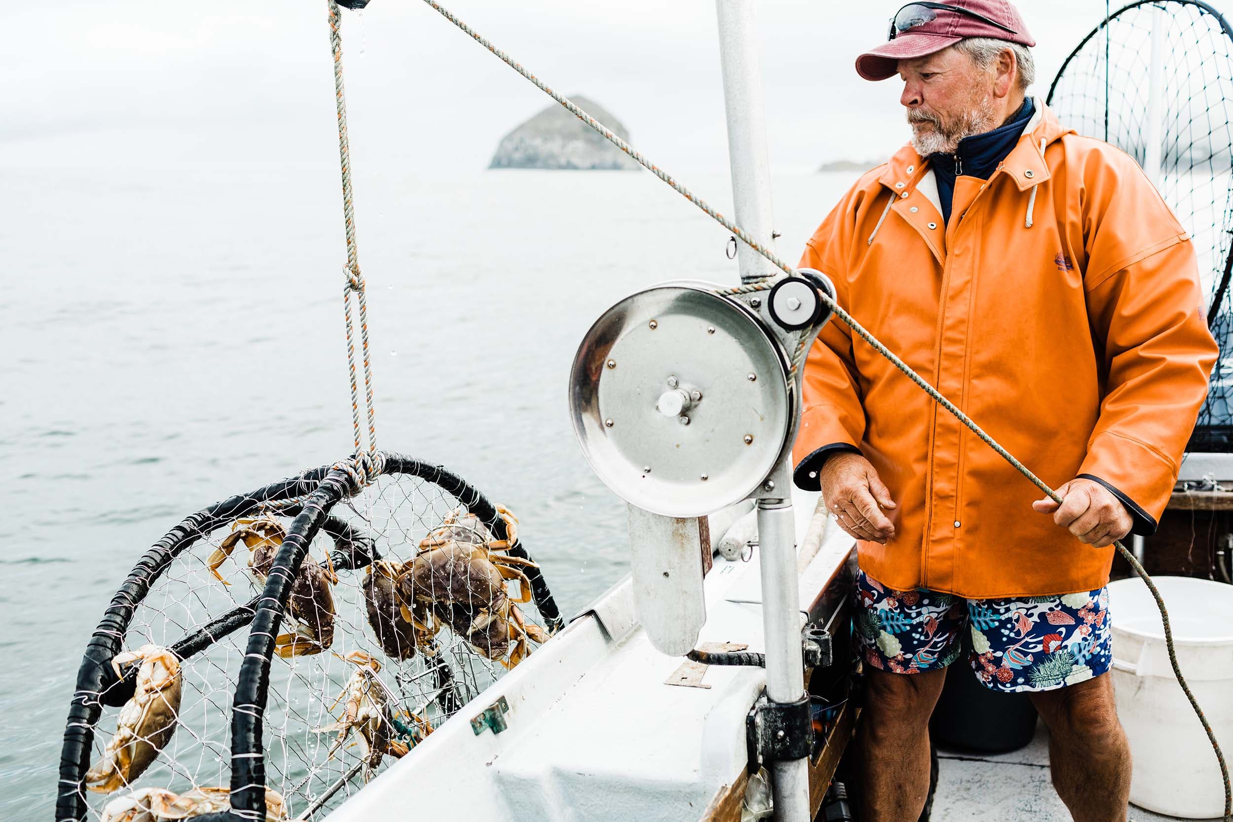 A man in an orange jacket and beach shorts on a fishing boats reeling in some crabs