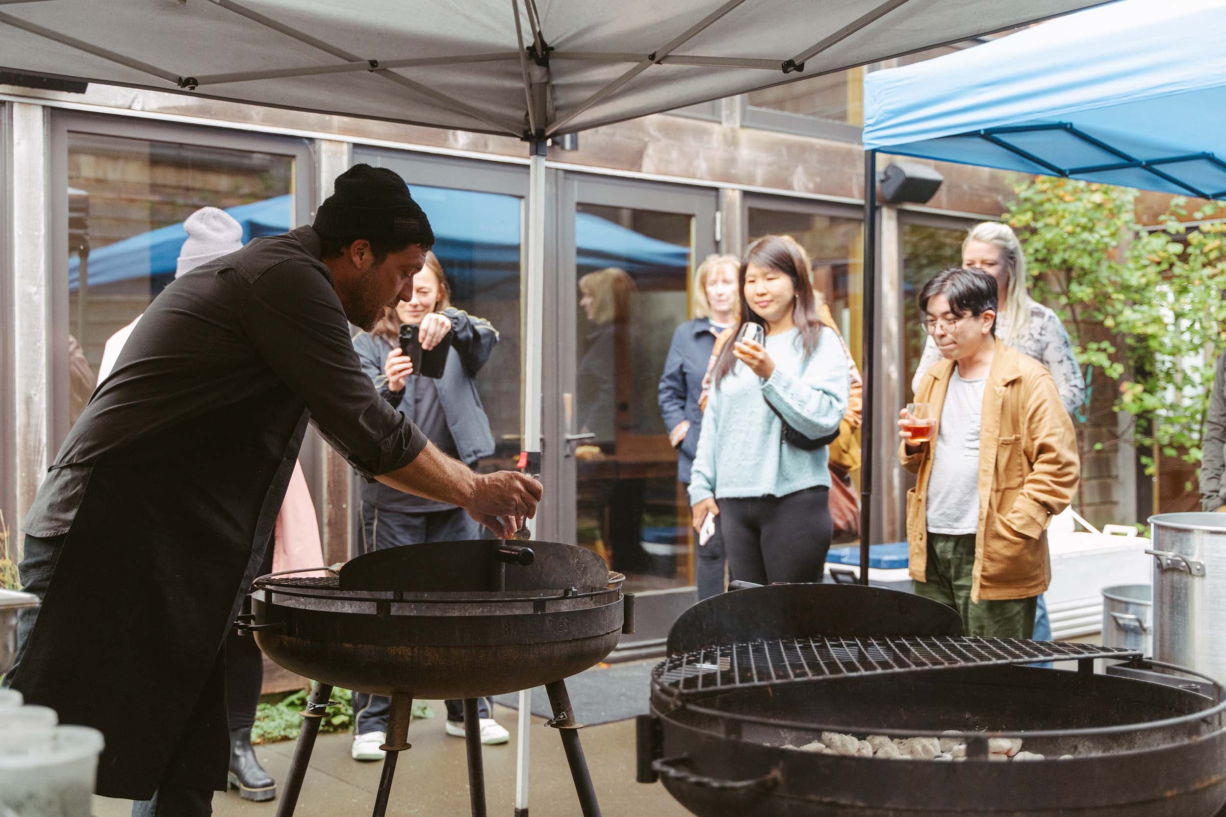 A man cooking on the BBQ while others are hanging out having drinks and taking photos of him at Headlands