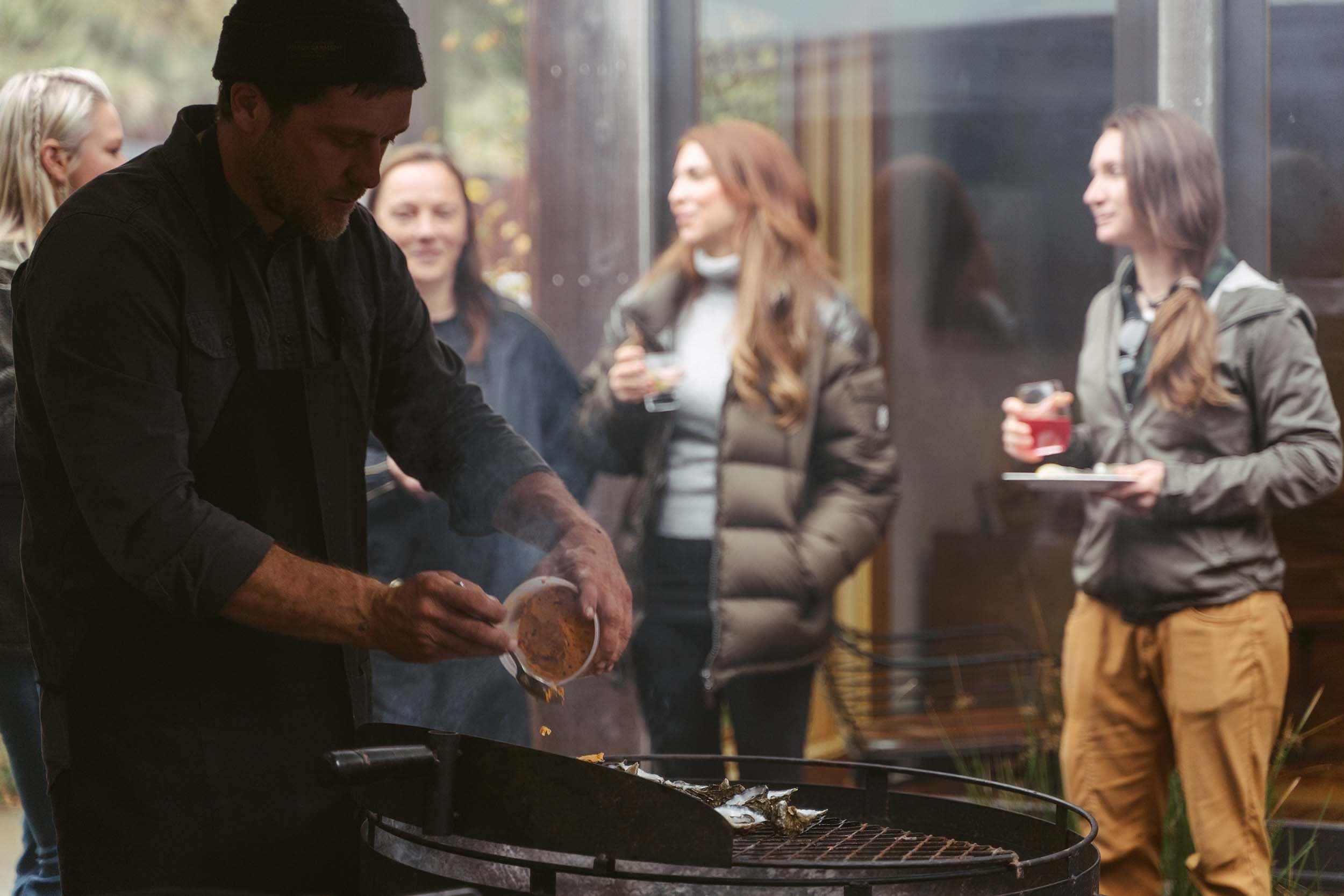 A man cooking on the BBQ while others are hanging out having drinks and a good time on the patio at Headlands