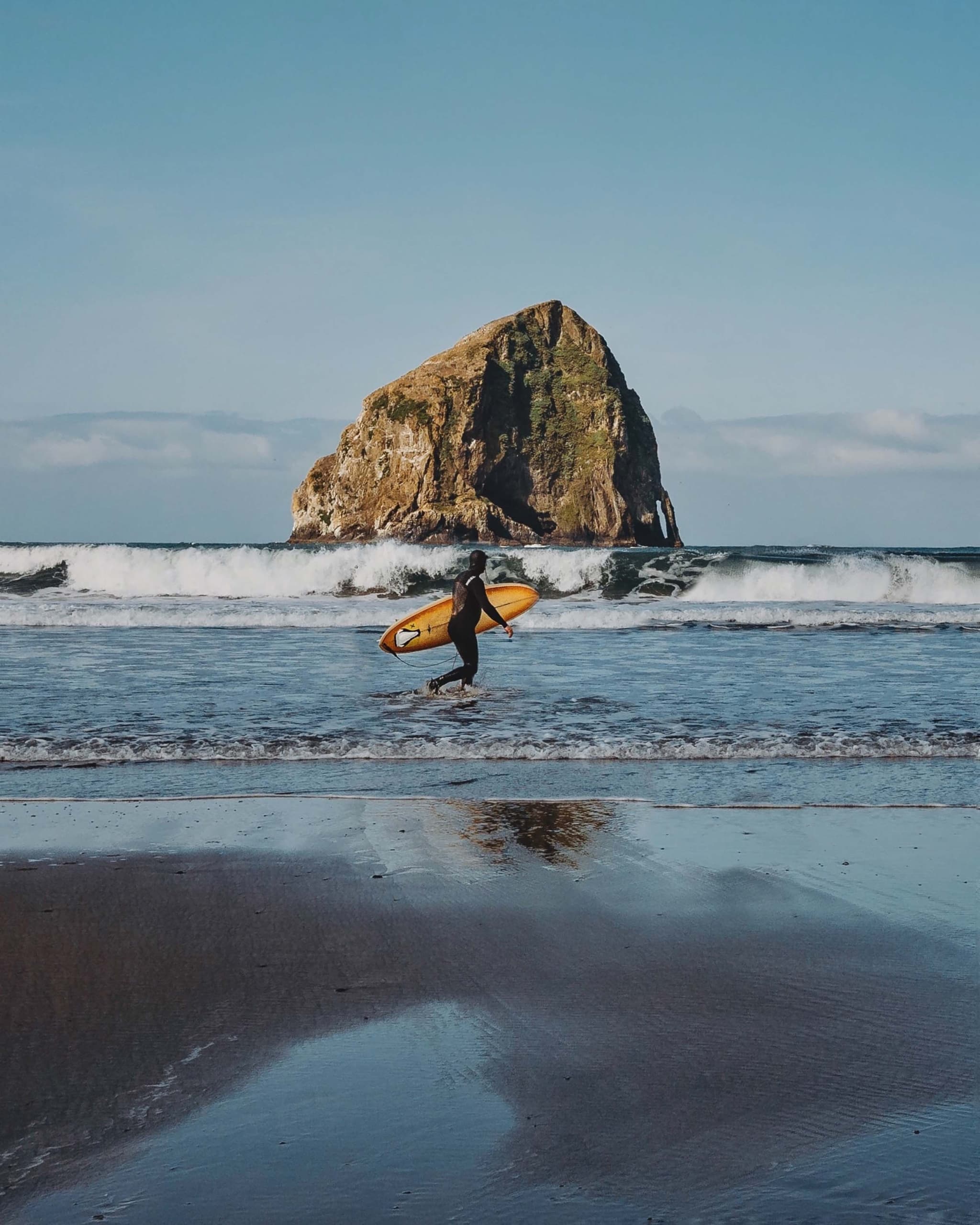 A sufer walking a long the beach in front of Haystack Rock